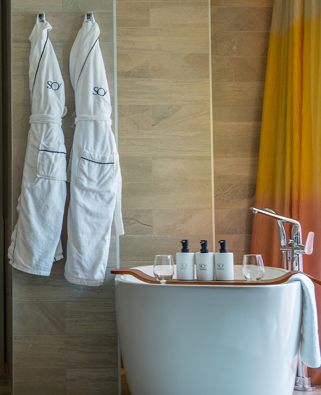 A wooden bath tray on a large bathtub with bath products and wine glasses. Next to it hangs two white 'SO/' brand bathrobes.