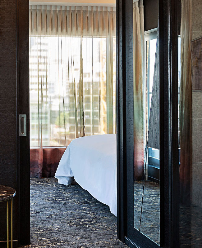 A doorway revelaing the corner of a bed and multi-coloured sheer curtains covering a window