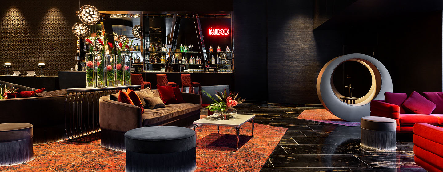 A bar with a neon 'MIXO' sign mounted on the wall with a seating area with couches, poufs and an 'O' shaped art piece