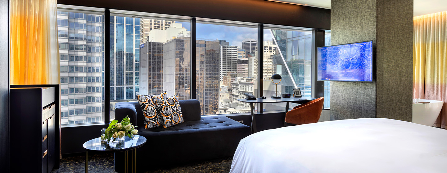 A large open plan hotel room with a bed, desk and blue couch facing a large window overlooking tall buildings