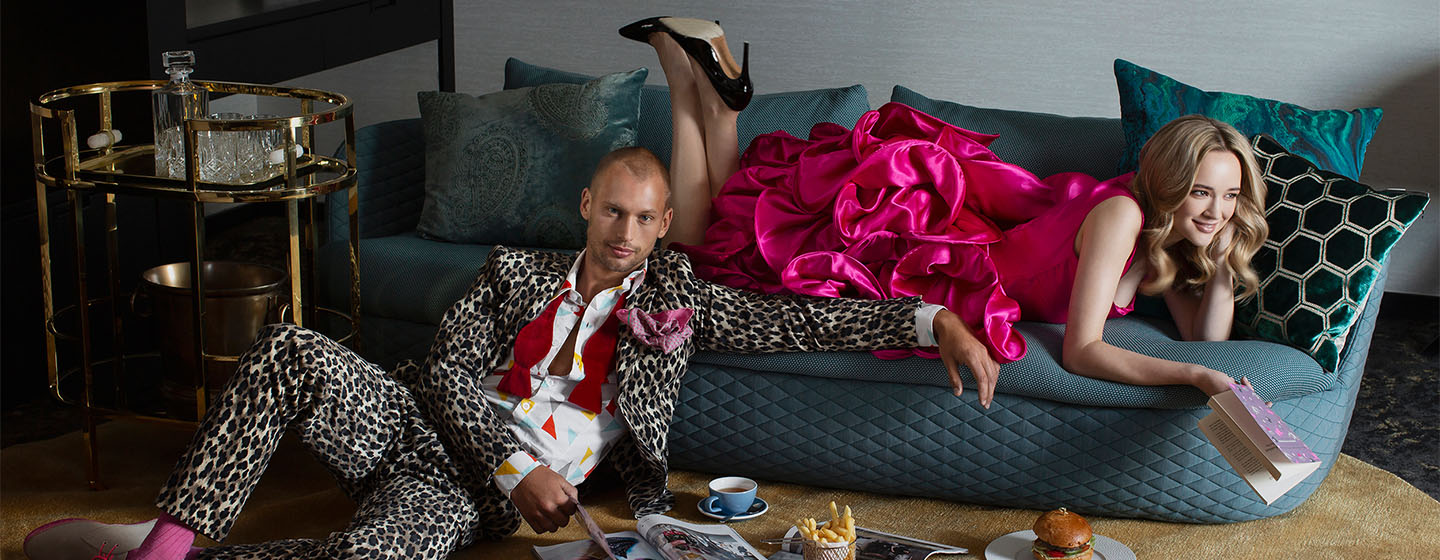 A woman in a pink frilled dress lays smiling on a couch whilst a man sits beside her on the floor in leopard pattern suit
