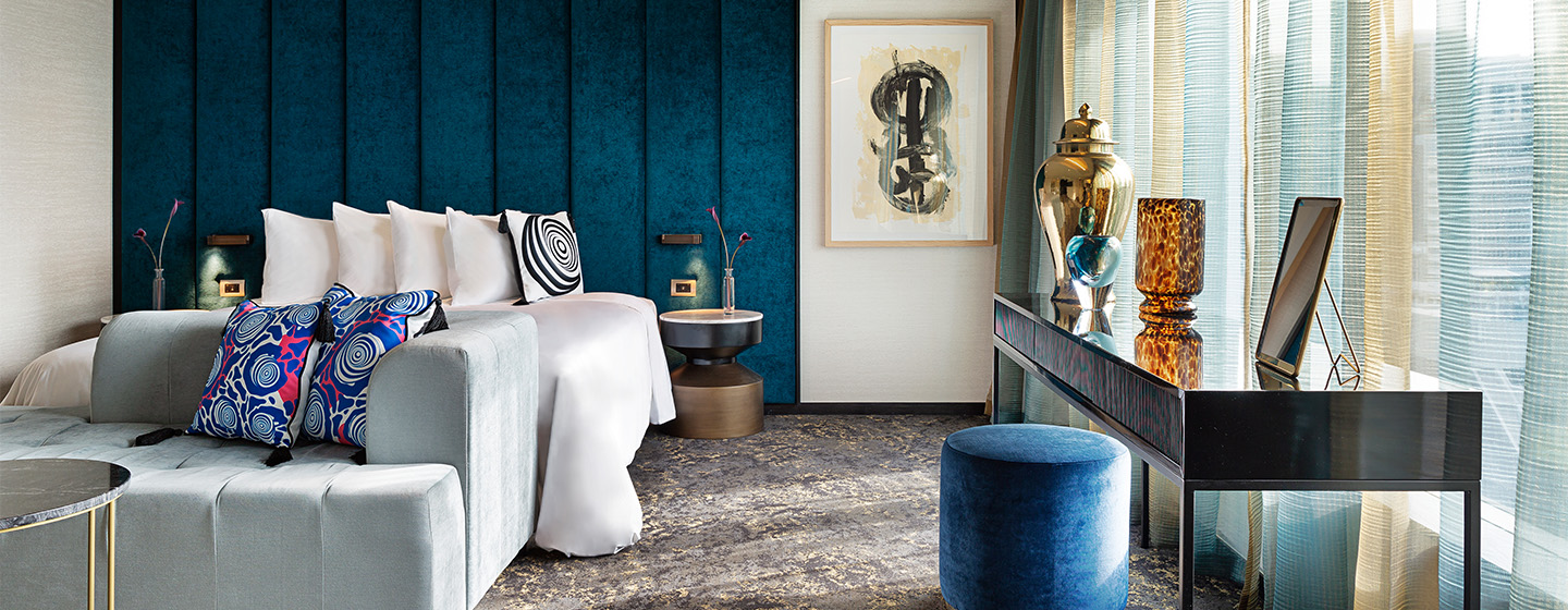 A modern hotel room with large bed, light blue couch and black cabinet topped with multiple ornaments.