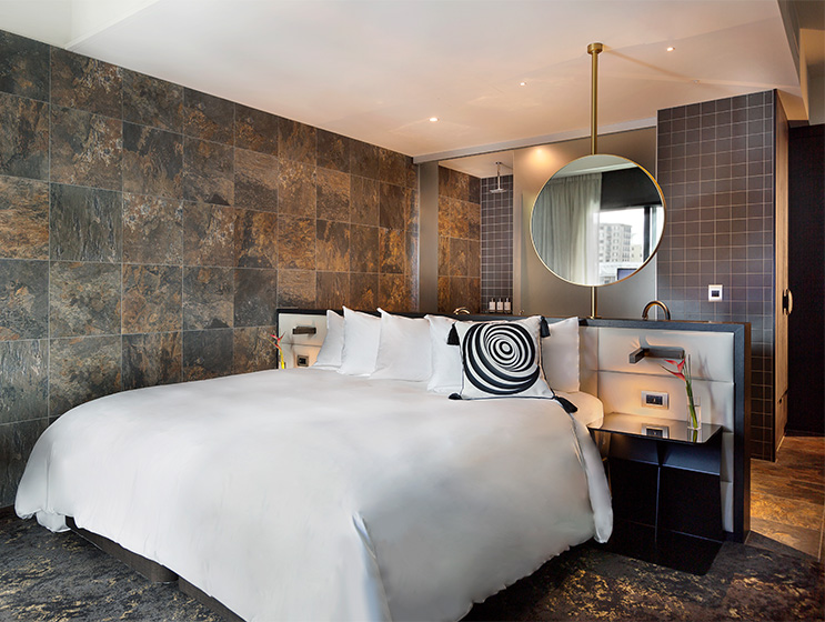 A double bed with side tables. Behind it a small wall sectioning of the tiled bathroom area.