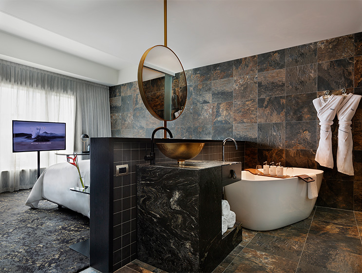 A small wall separates a bedroom and bathroom area with bathtub, gold sink and black counter top