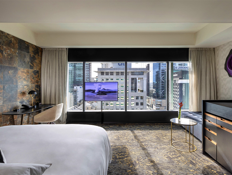 A large hotel room with a window with views of a city scape. A desk in on one side of the room, a cushioned bench on the other.