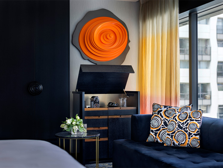 A dark blue couch next to a marbled coffee table and cabinet with a coffee machine and refreshments on it. A large orange emblem on the wall