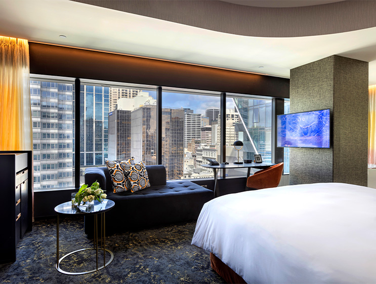 A large open plan hotel room with a bed, desk and blue couch facing a large window overlooking tall buildings
