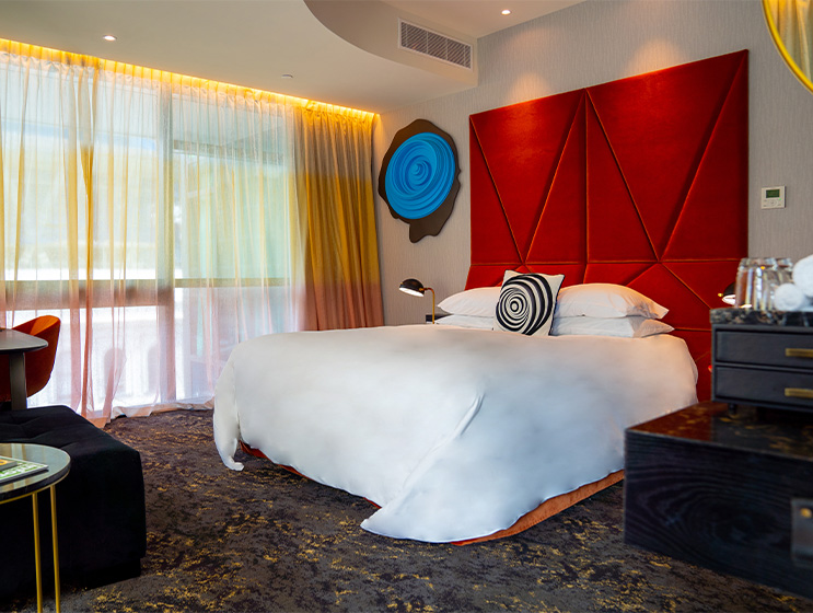 A hotel room with a large bed with a tall red headboard, a desk, and chest of drawers. On the wall a circular blue emblem.
