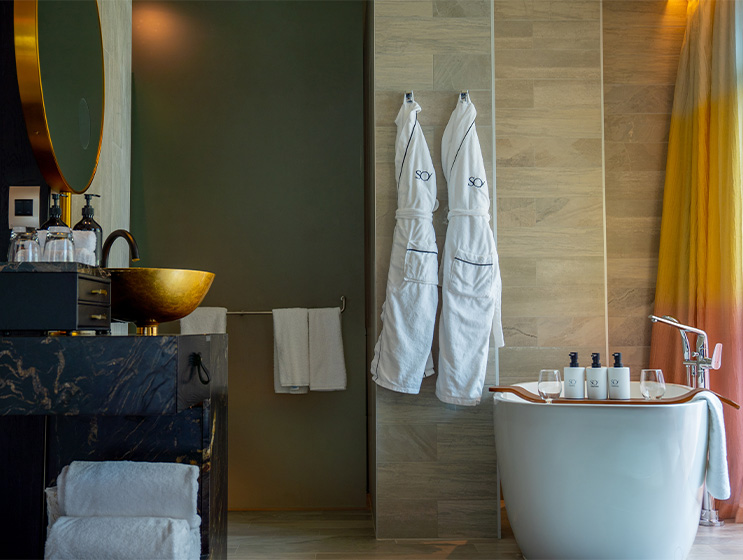 A bathroom with a large bathtub, next to it hangs two white 'SO/' brand bathrobes. To left left sits a gold sink and mirror on top of a black counter.