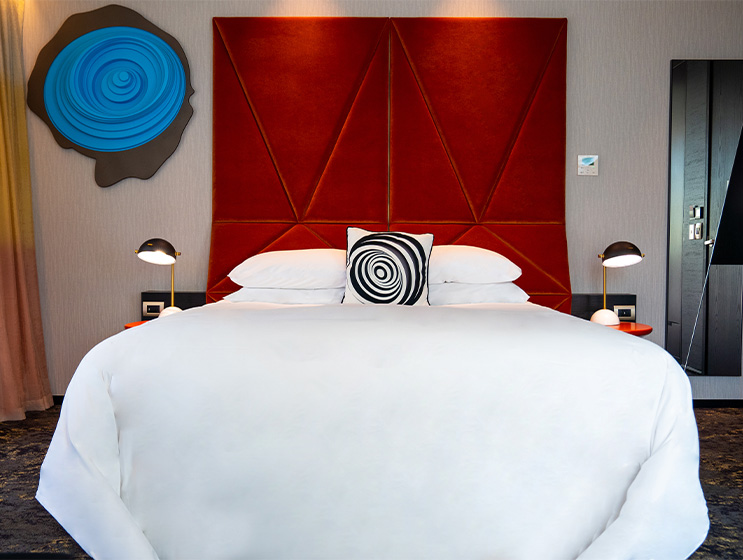 A large bed with a tall red headboard with a large blue emblem and mirrow mounted on the wall. There are bedside tables and lights on both sides.
