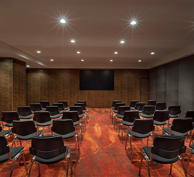A meeting room with burnt orange flooring and a flat screen TV mounted on the wall. Charis are layed out in rows facing the TV