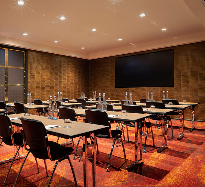 A small meeting room with tables and chairs layed out in rows facing a Flatscreen TV mounted on the walls.