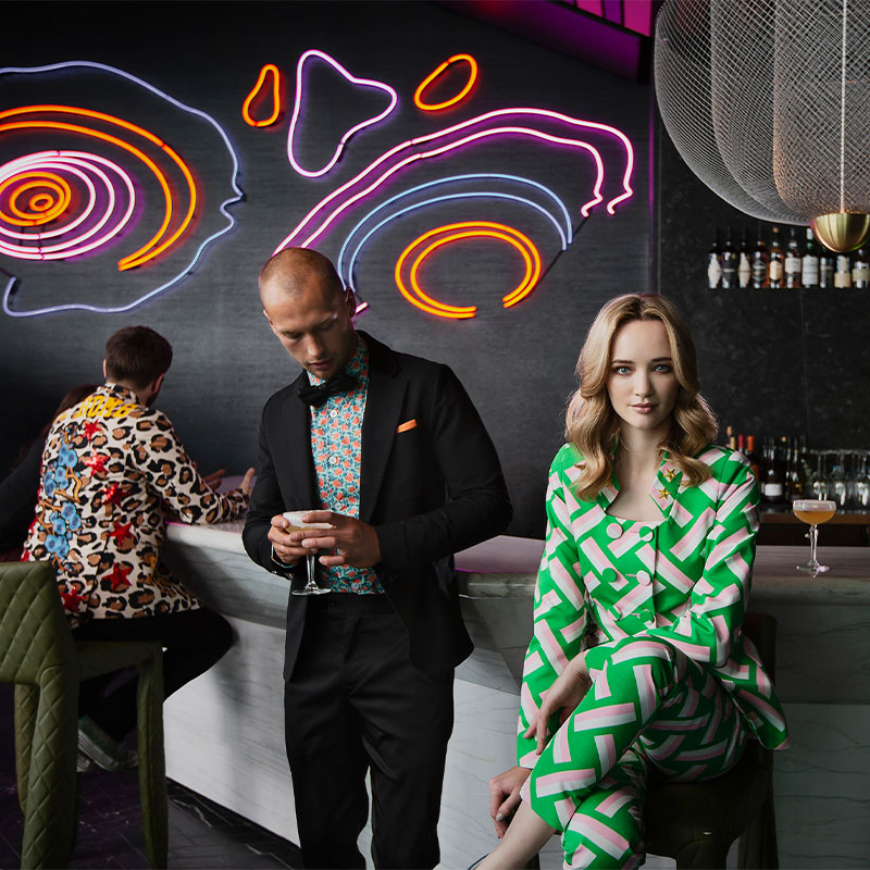 3 people standing and sitting by a bar. Behind the bar is a wall feature made of neon lights.