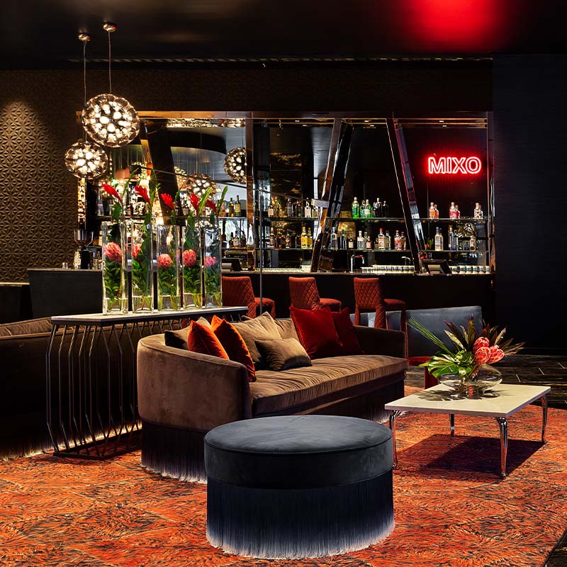 A bar with a neon 'MIXO' sign mounted on the wall with a seating area with couches, poufs and an 'O' shaped art piece
