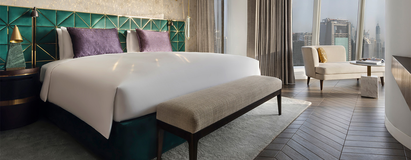 A king bed with green patterned head rest. A modern desk is situated next to floor to ceiling windows looking over the Dubai city scape.