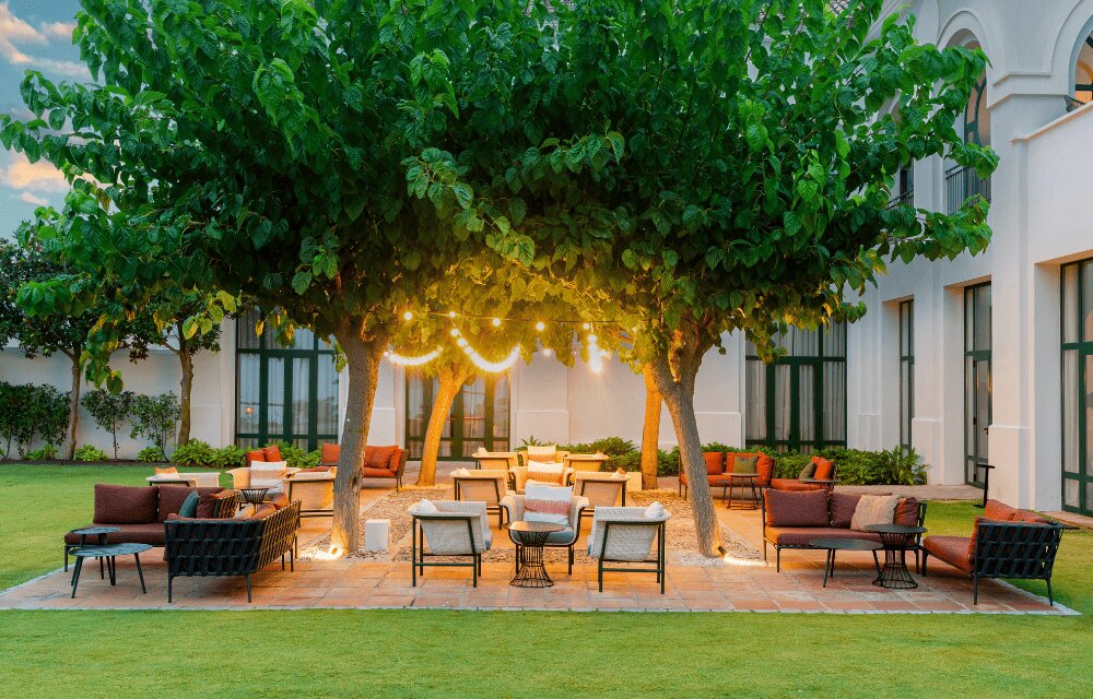 http://Outdoor%20patio%20scene%20with%20couches%20and%20chairs%20under%20two%20large%20trees,%20adorned%20with%20string%20lights.%20The%20setting%20is%20cozy%20and%20inviting,%20located%20beside%20a%20building.