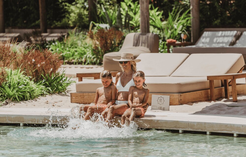 http://A%20woman%20in%20a%20sun%20hat%20and%20white%20swimsuit%20sits%20by%20a%20pool%20with%20two%20young%20children,%20splashing%20water.%20The%20scene%20is%20relaxed%20with%20a%20tropical,%20sunny%20vibe.