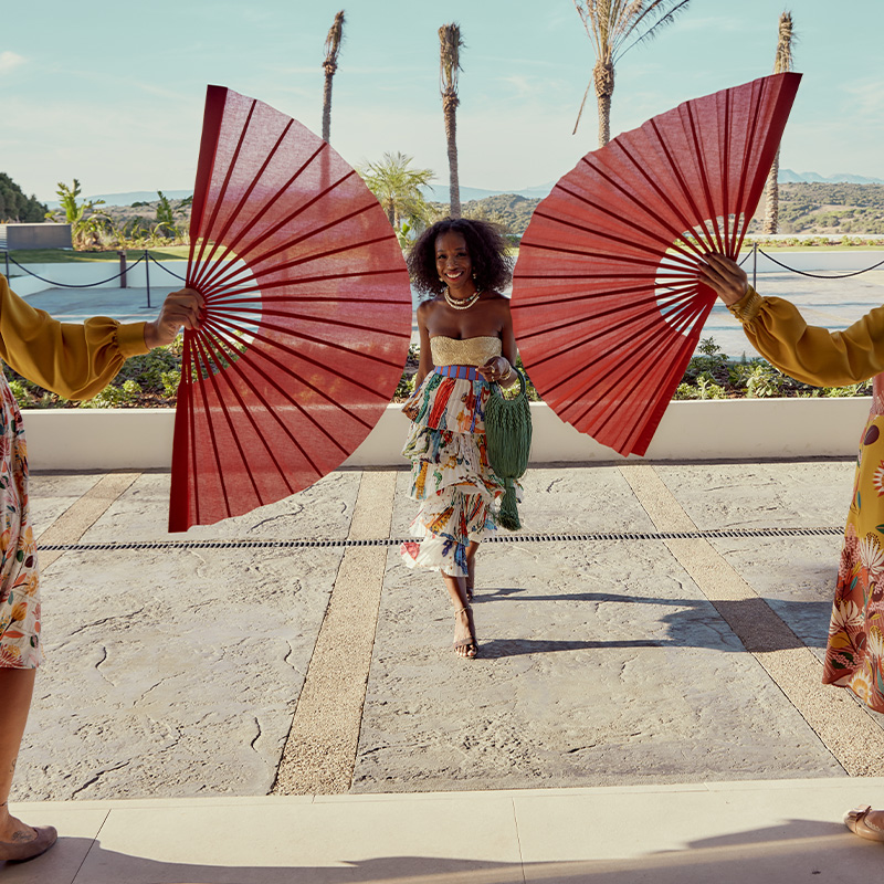 A woman in a flowery dress about to walk through two people holding large red fans