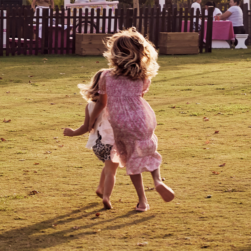 A little girl in a pink dress plays with another little girl on a grass field