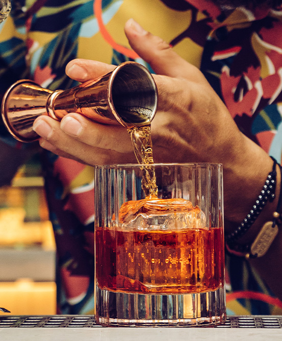A man in a colourful shirt pours a shot into a whisky glass over a large ice cube