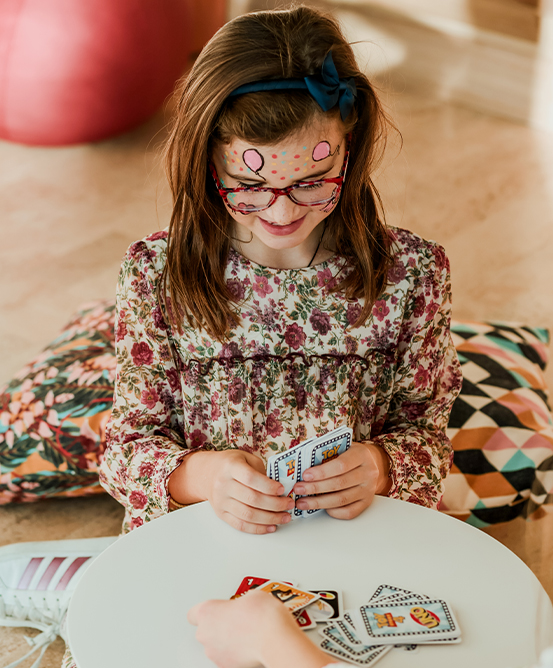 A little girl in a flowery dress and her face painted plays uno with someone out of frame