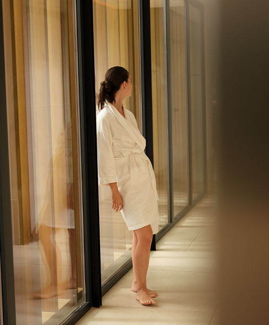 A woman looking down a corridor in a white robe leaning against floor to ceiling windows