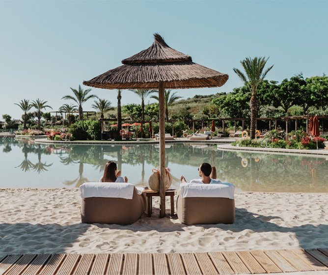 A man and woman sitting on loungers on a sandy beach underneath a wicker parasol