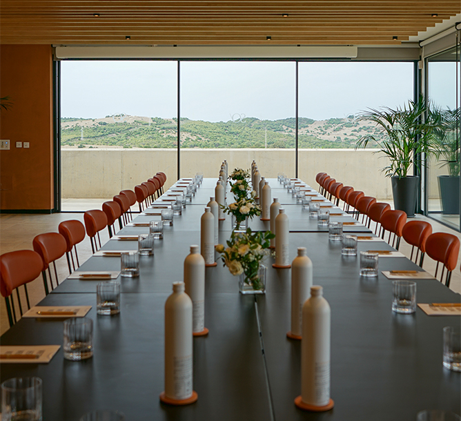 A meeting room with a long meeting table equipped with stationary and bottles of water and full to ceiling windows with views of a mountain