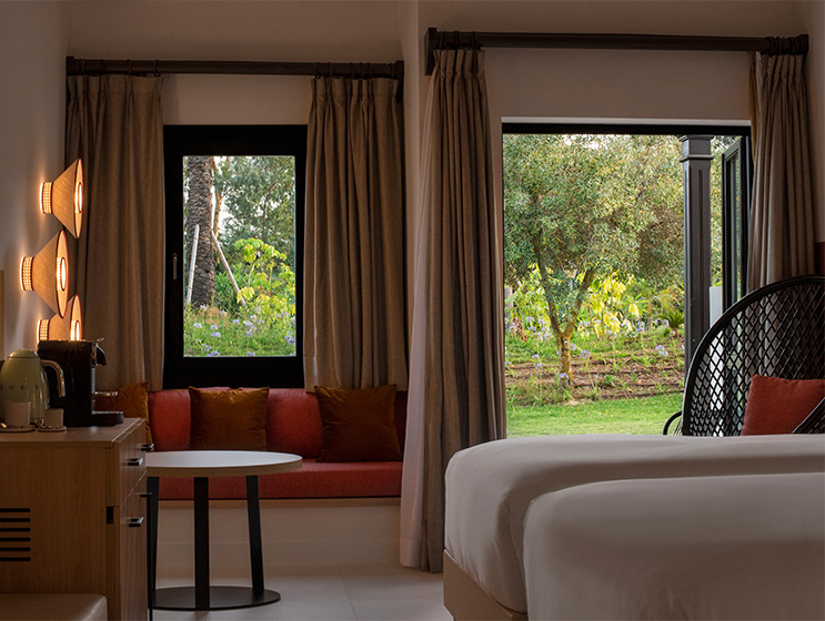 A dimly lit hotel bedroom with an open door leading onto a large green garden area.