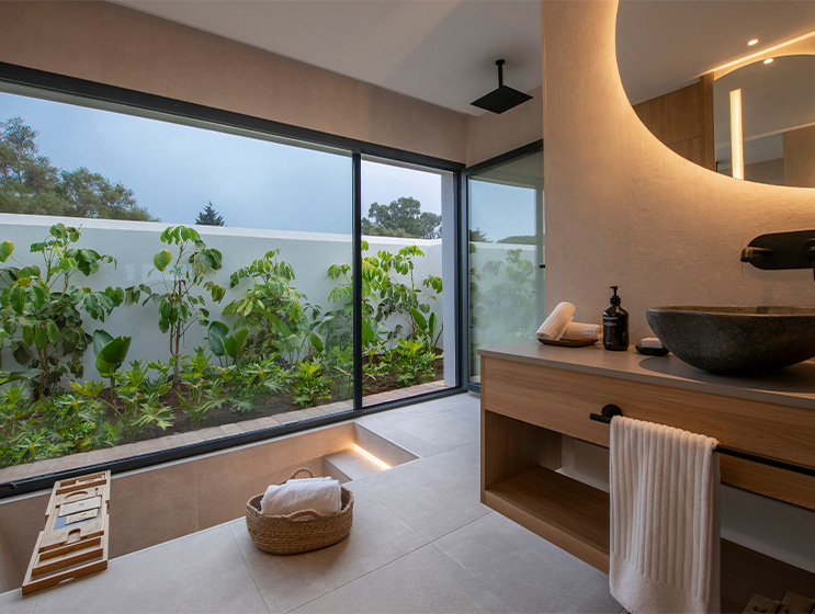 A large open bathroom with a deep bath, rain shower and large window displays a garden area