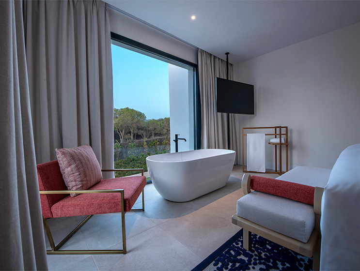 A bathtub sits in front a window in a hotel room. There's also a chair, cushioned bench and TV hanging from the ceiling