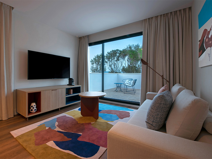 A lounge area with a large couch facing a mounted flat screen TV. A large window looks out onto a terrace