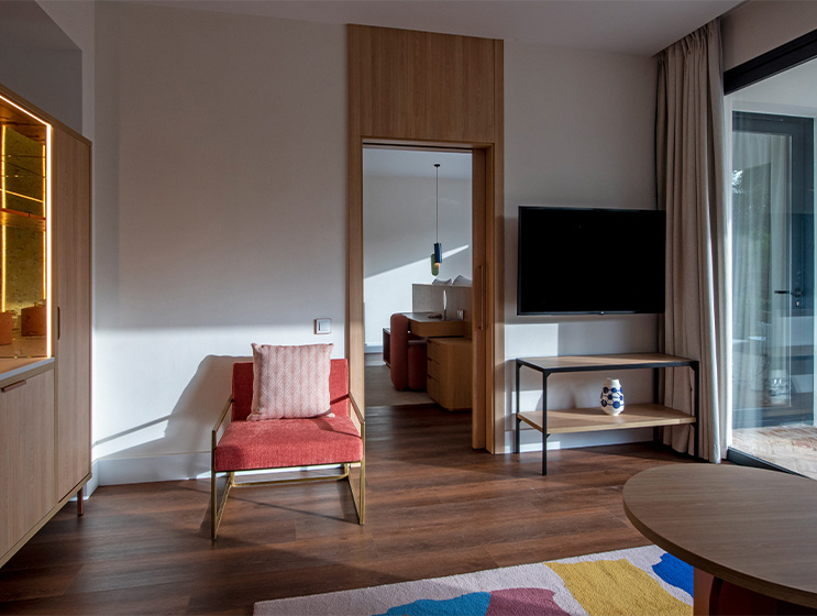 A lounge area with TV, armchair and large window leads onto a bedroom area