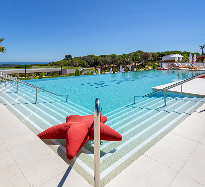 A large pool with clear blue water and a backdrop of trees and the sea. A starfish shape float sits on the stairs of the pool