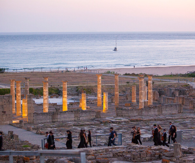 A group walk by brick ruins whilst in the distance a beach and a boat sailing on the water