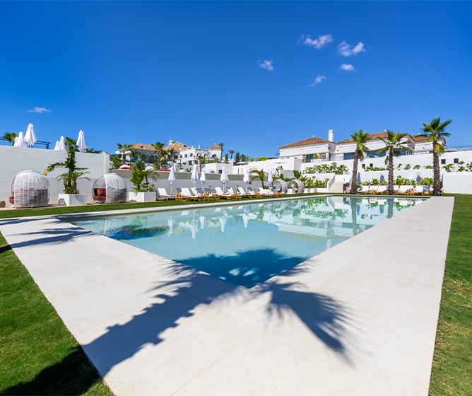 A pool surrounded by loungers, circular booths and palm trees under a clear sky. In the distance are white andalusian style buildings