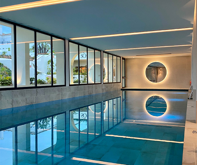 A large indoor pool. At one end is an illuminated circular mirror and large windows cover one side of the wall. 