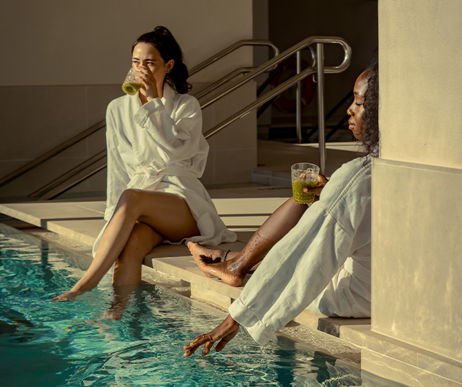 Two women in bathrobes lounging on the edge of an indoor pool drinking green bevarages.