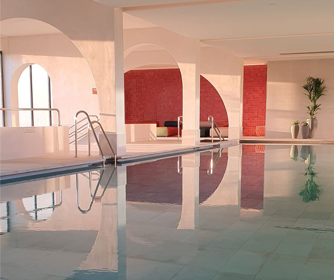 A large indoor pool with the refflection from a red tiled wall reflecting a pink glow over the white space