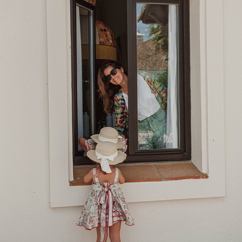 A little girl attempts to climb out of a window to join another little girl whilst her mother looks on