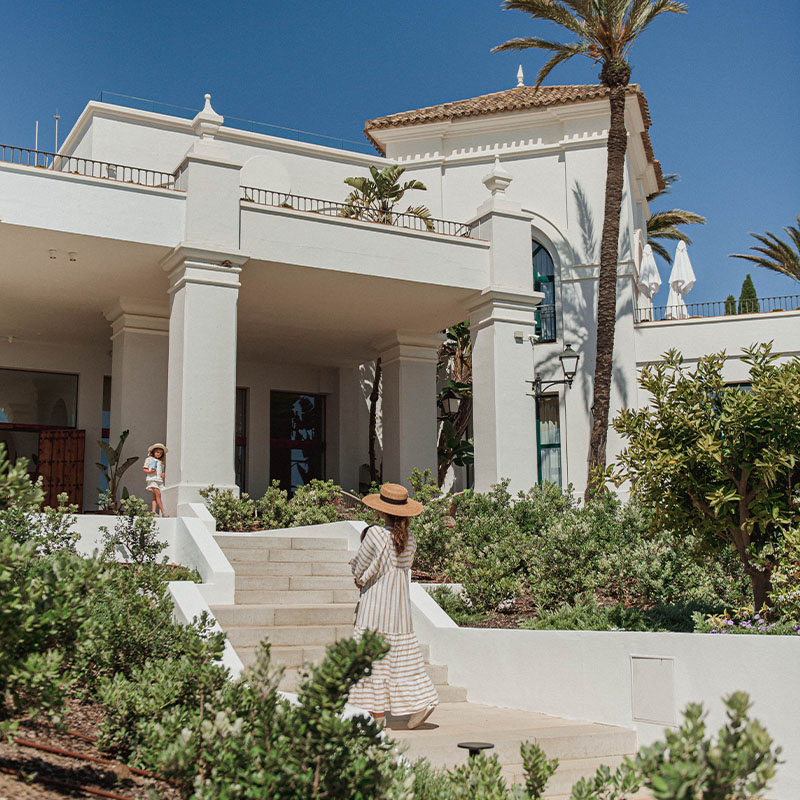 A woman in a summer dress walks up the steps towards a large white andalusian style buidling