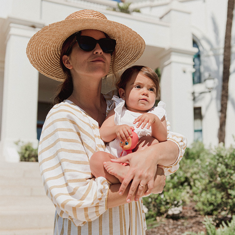 A mother in a summer dress and straw hat, holds her baby looking out into the distance