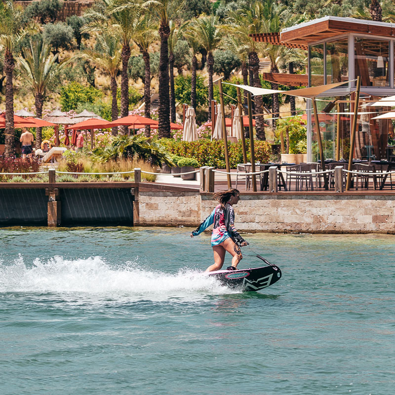 A women riding an electric surfboard on clear blue water. Behind her large palm trees, parasols and seating area