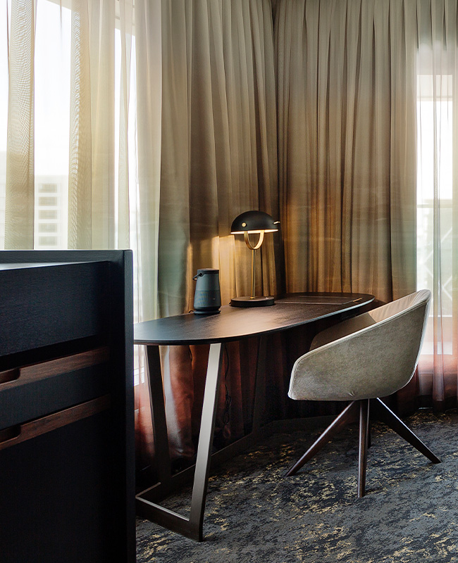 A dark wooden desk sits in the corner of a room with a circular desk chair. The windows are covered by sheer curtains.