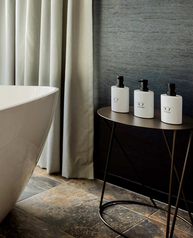 Three SO/ branded bath products sit on a small table next to a white bath tub