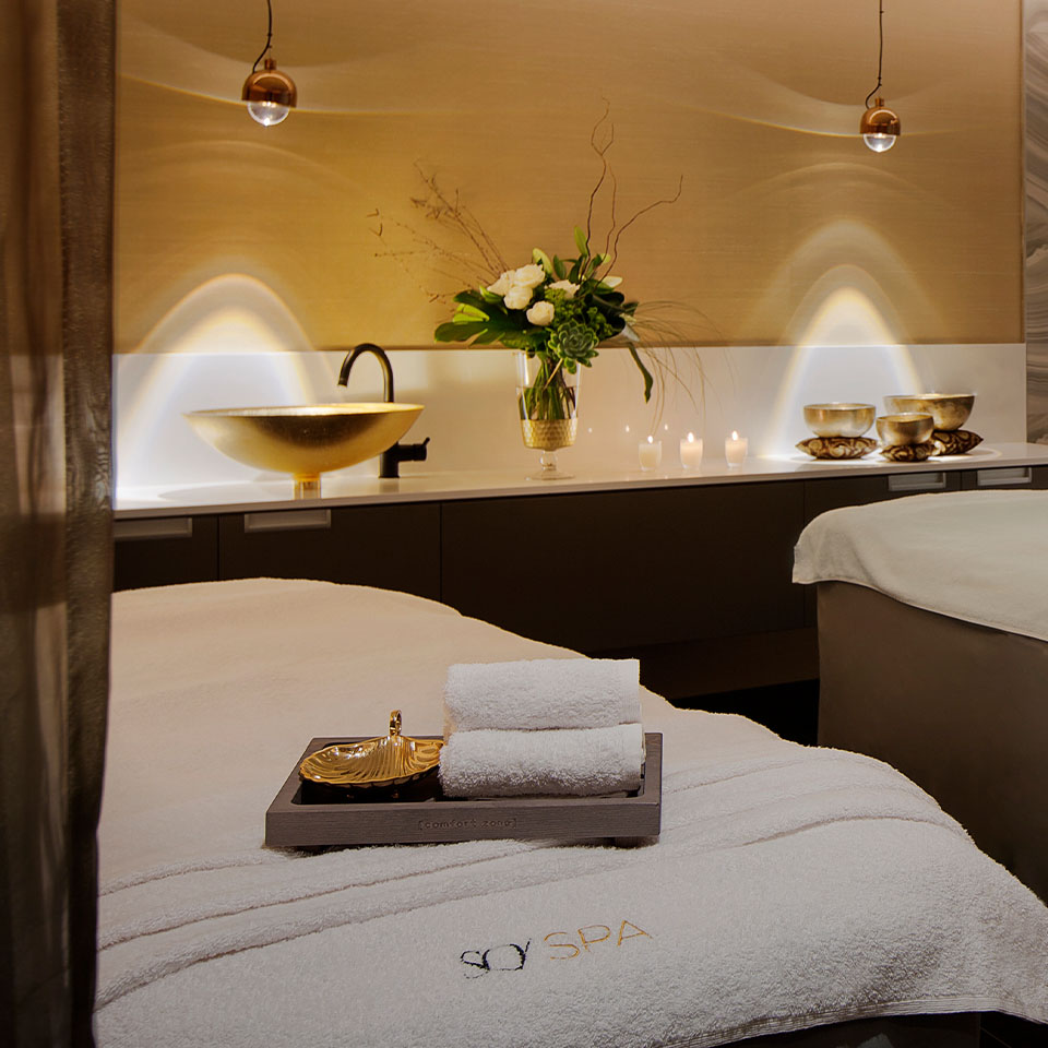 A spa treatment room with two massage tables. A gold sink, singing bowls, candles and flowers sit on a work surface behind them.