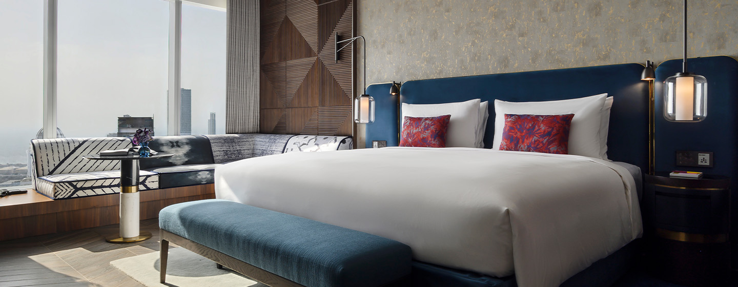 A large bed with blue velvet head rest. A modern sofa-bench is situated next to floor-to-ceiling windows looking over the Dubai.