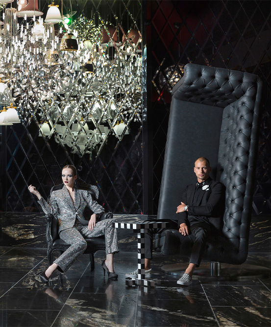 A man sitting on a upturned couch and a woman posing next to him in an armchair. A large complex chandelier hangs low beside them