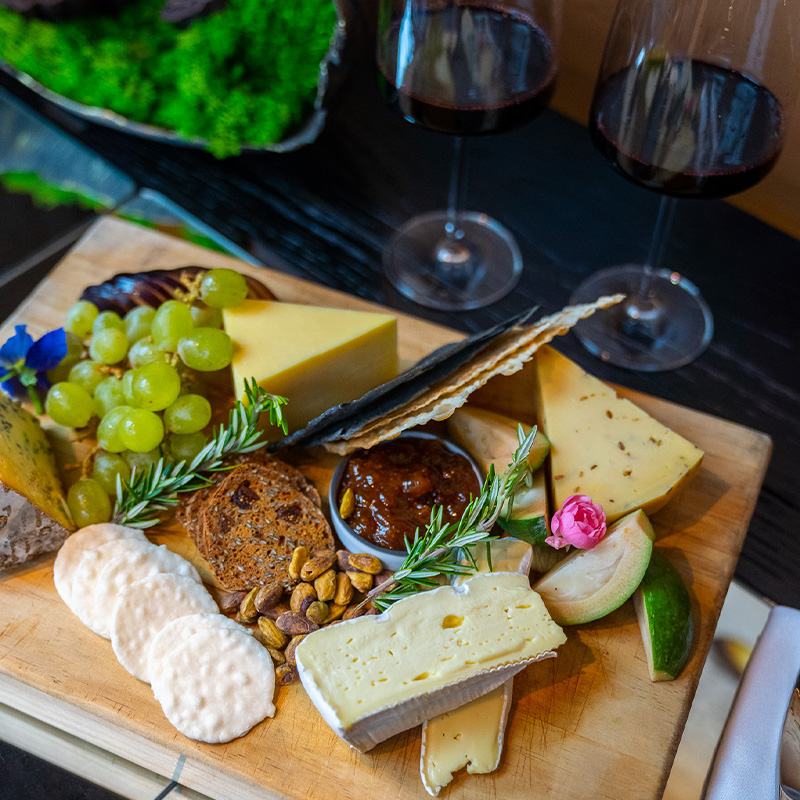 A wooden board filled with grapes and a vareity of cheese and biscuits next to two wine glasses.