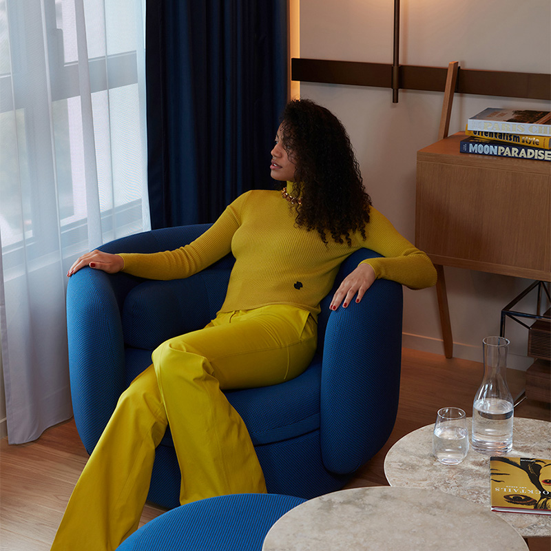 A women in a yellow outfit sitting on a blue armchair with glasses of water next to her on marble coffee tables